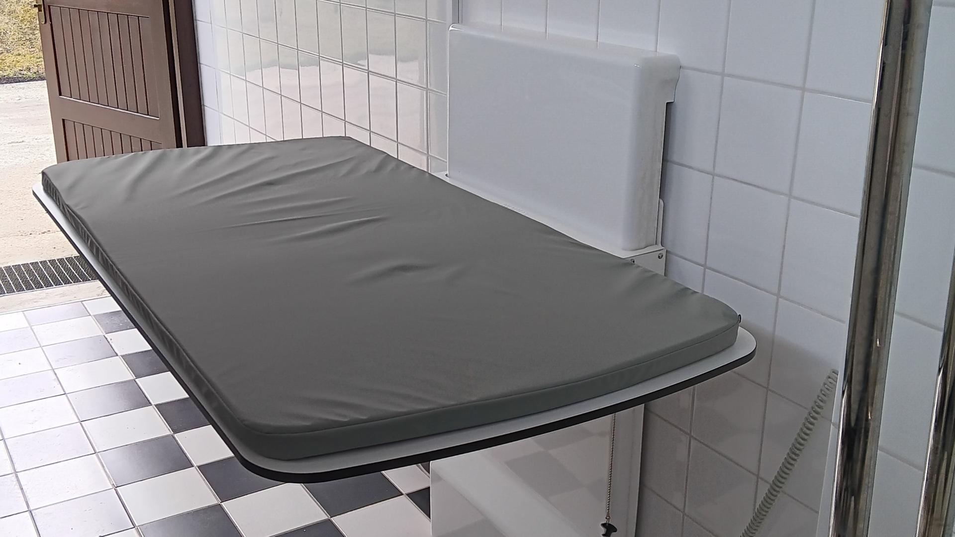 We wnętrzu komfortki rozłożna leżanka sterowana elektrycznie, na niej ciemnoszary materac. W głębi otwarte brązowe drzwi wejściowe do komfortki, która jest jednocześnie toaletą dla osób z niepełnosprawnością. Na ścianie błyszczą białe kafelki, a na podłodze biało-czarna terakota.