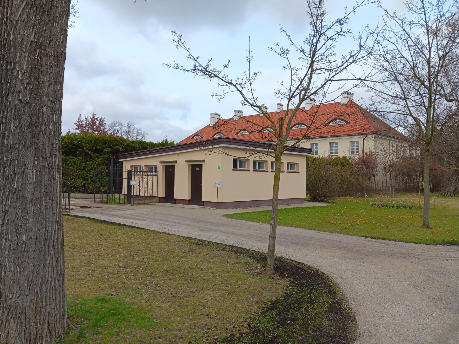Kremowy parterowy budynek, w którym znajdują się toalety, w tym dla osób z niepełnosprawnością i komfortka. Dobrze widoczny z daleka. Stoi przy asfaltowej alei i jest oznakowany. Za nim piętrowy budynek z dachem z czerwonej cegły.