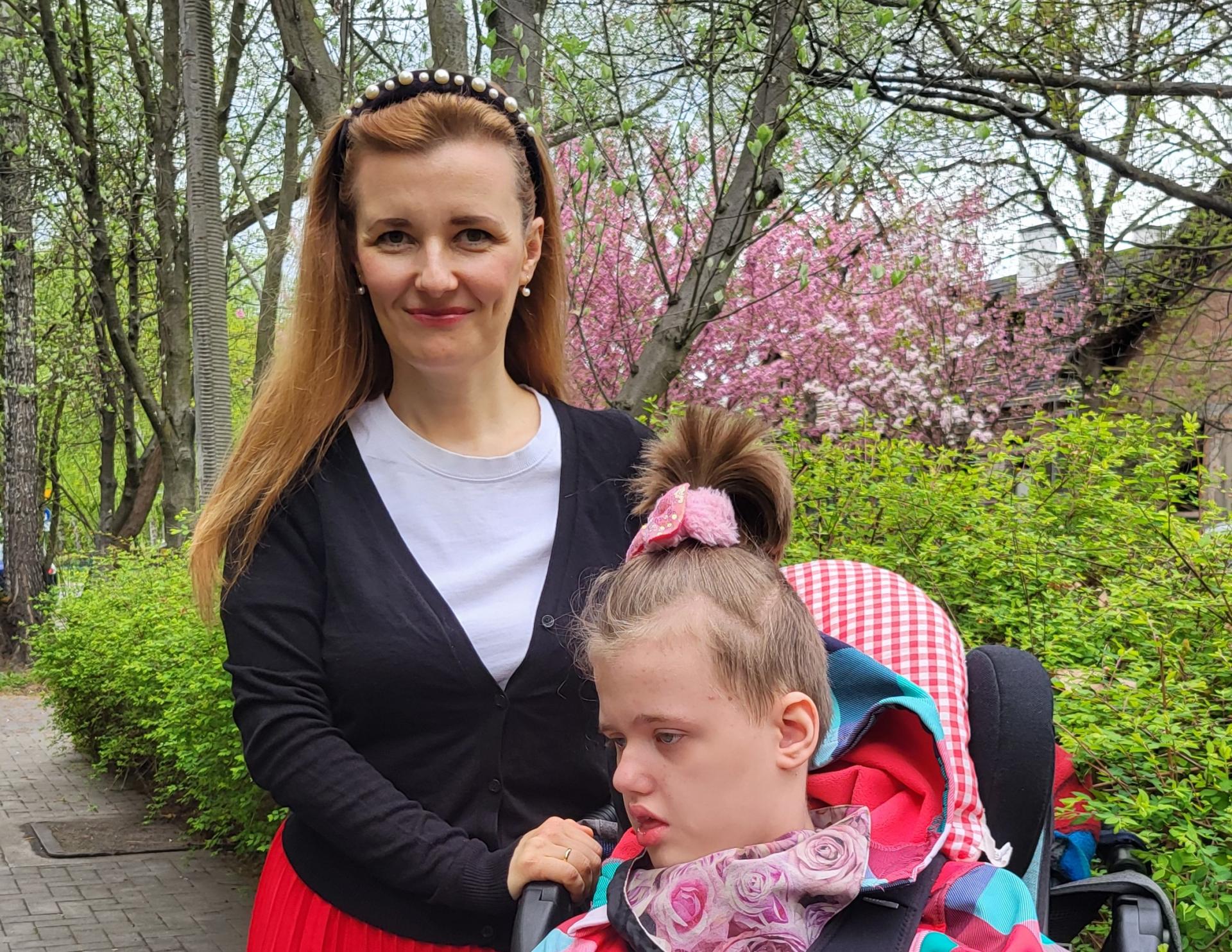 Na tle majowej zieleni i kwitnących drzew stoi kobieta w białej bluzce, czarnym sweterku i czerwonej spódnicy, trzymając przed sobą wózek z ok. 10-letnią dziewczynką w kolorowej kurtce i z kucykiem na czubku głowy/