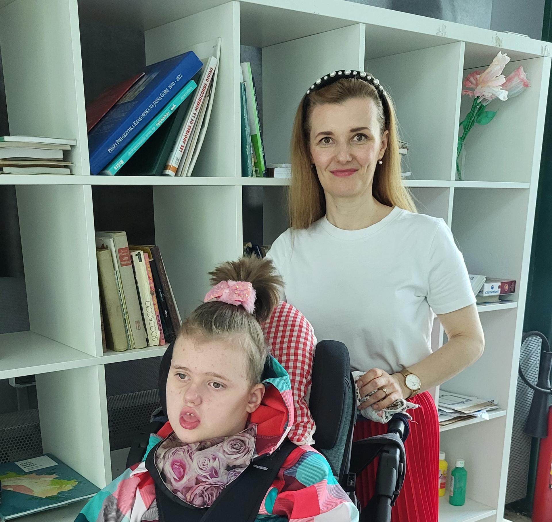 Kobieta w białej bluzce i czerwonej spódnicy stoi za wózkiem, w którym siedzi 10-letnia dziewczynka w kolorowej kurtce i w kucyku na czubku głowy. W tle biały regał z różnymi publikacjami, papierami i farbami.