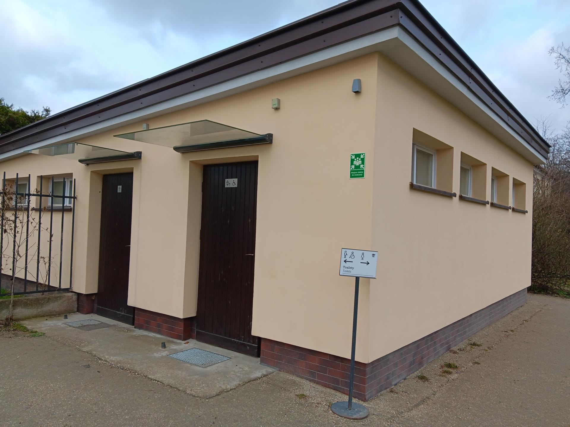 Kremowy parterowy budynek, w którym znajdują się toalety, w tym dla osób z niepełnosprawnością i komfortka. Zbliżenie na dwoje drzwi, bezprogowe wejście i oznaczenie w systemie Totupoint.