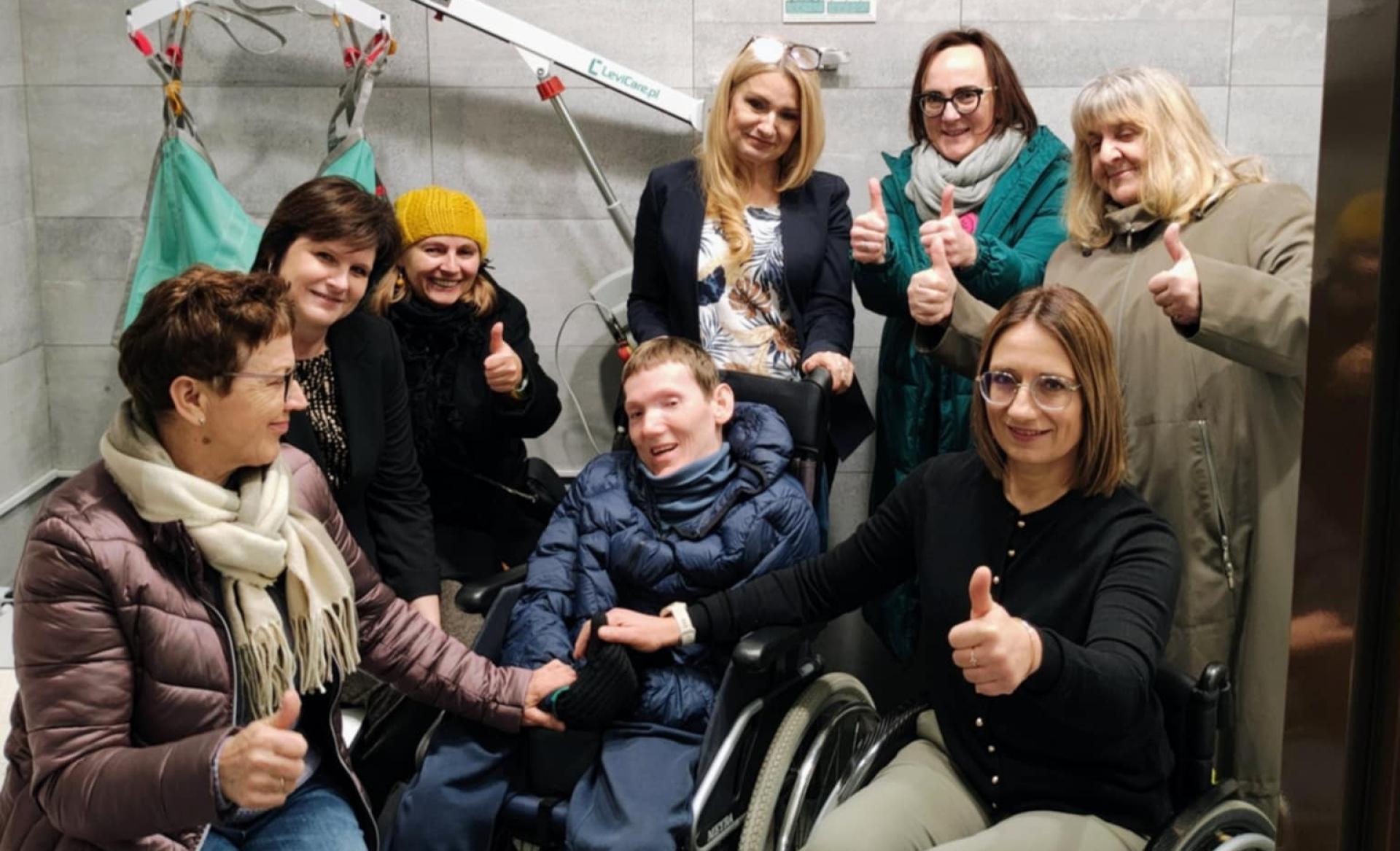 Na zdjęciu jest grupa osób pokazująca kciuk do góry. Pośrodku jest uśmiechnięty mężczyzna siedzący na wózku.