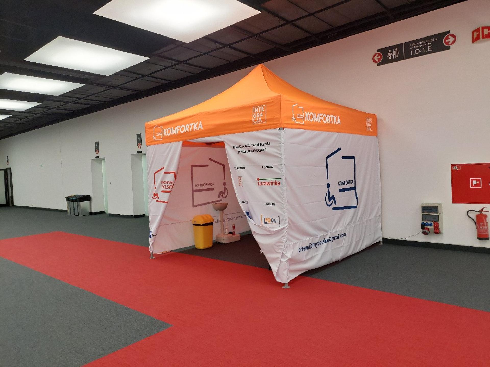 Komfortka plenerowa we wnętrzu - przestronny odpowiednio wyposażony namiot, tuż obok toalet, ustawiony w szerokim korytarzu