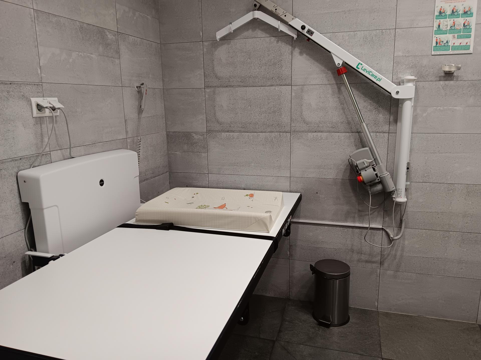 Pomieszczenie z leżanką, regulowaną elektrycznie, oraz podnośnikiem przymocowanym do ściany, także regulowanym elektrycznie